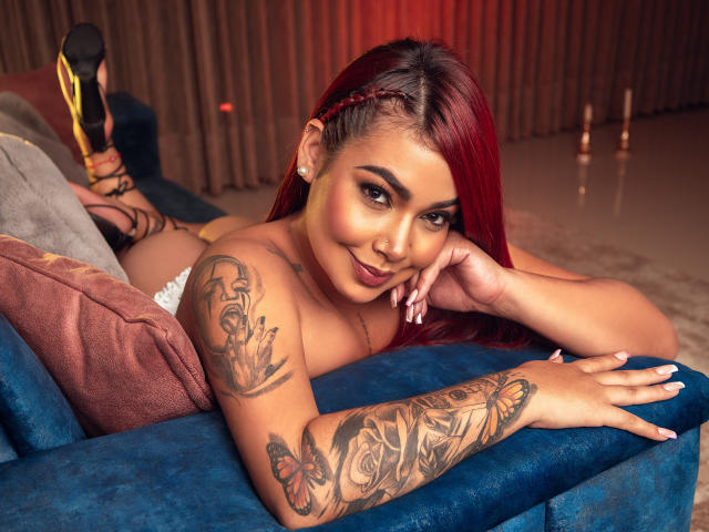 ValeryJhonston, avec des cheveux roux, allongée sur un canapé bleu, montrant des tatouages sur les bras, portant des talons hauts, souriant à la caméra, une main sur la joue, ayant l'air séduisante, sans vêtements..