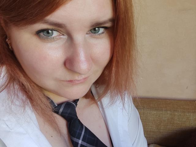 MinistraX has pale skin, green eyes, and red hair, wearing a white shirt with a black plaid tie, and a small nose ring, with a subtle, confident smirk.