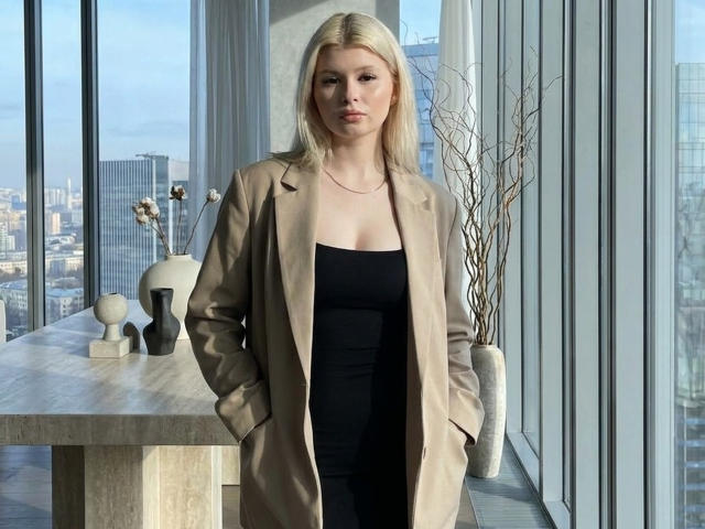 Delphine Hambleton, con cabello rubio, vestida con un abrigo beige sobre un vestido negro, con las manos en los bolsillos, de pie junto a una ventana con vistas a la ciudad, con una expresión seria, con los senos visibles debajo del vestido..