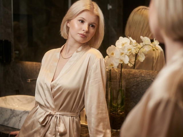 Delphine Hambleton, con i capelli biondi tagliati corti, indossa un morbido accappatoio beige, mentre si guarda allo specchio, mostrando la sua pelle liscia e chiara e i suoi piccoli seni, con un vaso di orchidee bianche..