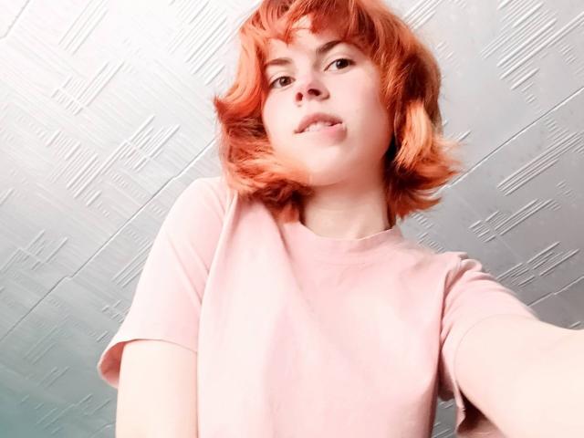 MireilleSpotwood with orange hair, wearing a pink shirt, taking a selfie, looking slightly down with a relaxed expression.