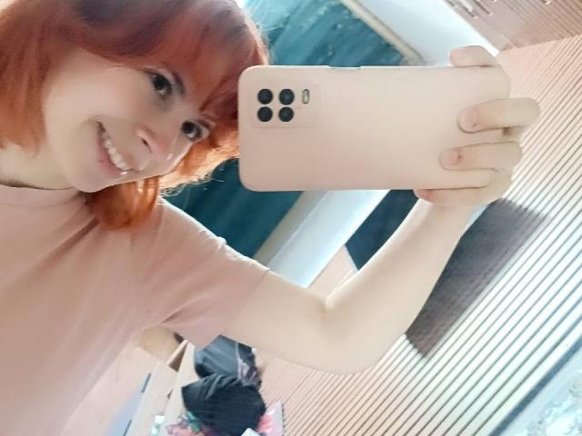 Mireille Spotwood, con i capelli rossi, sorridente, che tiene in mano un telefono rosa, sta scattando un selfie, indossa una maglietta rosa, mostra il suo braccio e la sua mano, con i seni parzialmente visibili..