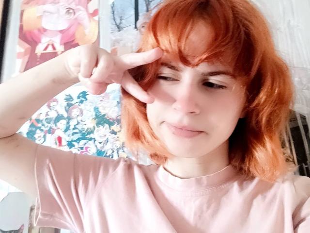 Mireille Spotwood, con i capelli rossi, che fa un gesto di pace, ammiccando, indossa una maglietta rosa, ha un sorriso carino, pelle chiara e un viso leggermente paffuto..