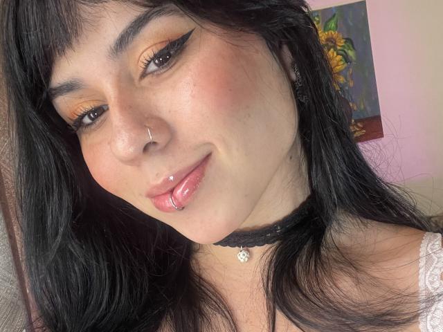 GaiaJoness tem cabelo preto, um piercing no nariz e um piercing no lábio. Ela está usando delineador laranja, um colar preto e uma blusa branca de renda. Seus lábios estão brilhantes e rosados..