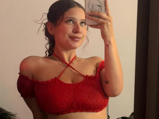 ValeryScot with curly brown hair, red off-shoulder crop top with crisscross straps, small boobs, holding phone, smiling, light skin, tattoo on right arm, reflection in mirror.