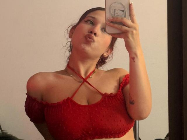 ValeryScot with pouty lips, wearing a red off-shoulder top with criss-cross straps, holding a phone in front of a mirror, showing small tits and tattoos on her arm.