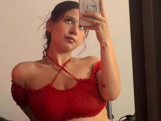 ValeryScot with wavy brown hair, red off-shoulder crop top showing her small tits, holding phone, subtle tattoo on right arm, pouty expression, reflective mirror selfie.