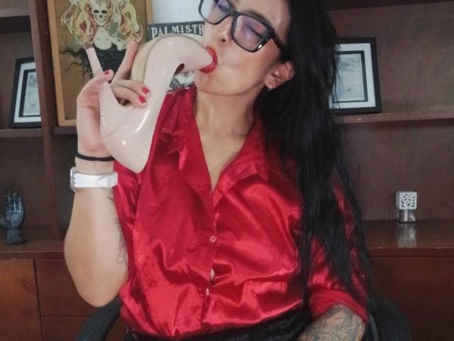 ZarayFeet with long black hair, glasses, and red silk shirt, sucking on a pink phone's curved receiver, showing tattooed arm and white wristwatch.