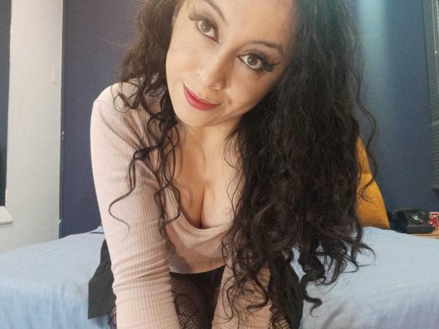 KendraSimons with curly black hair, red lipstick, and a pink ribbed top, leaning forward, showing her large boobs, and smiling at the camera.