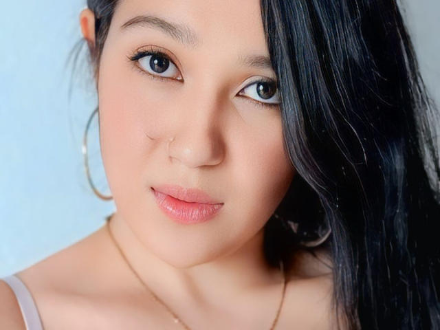 LuxyLeim's face with glossy pink lips, dark straight hair covering one eye, hoop earrings, subtle makeup, and a gold necklace, staring directly at the camera with a slight smile.