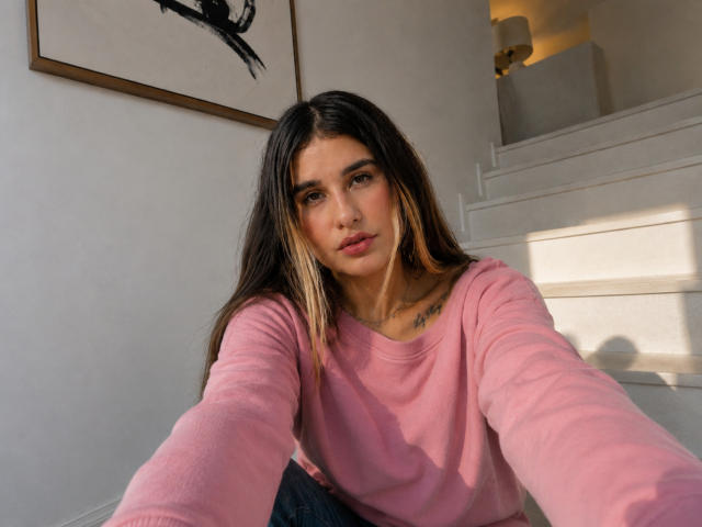 KatieDesirX, com cabelos castanhos longos, um suéter rosa e uma tatuagem no peito, tirando uma selfie, mostrando seu rosto e a boca ligeiramente aberta, em um quarto iluminado pelo sol..