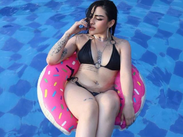 LilitthCorper with black bikini, pink donut float, tattoos, and piercings, sitting in blue pool, biting finger, looking down, legs crossed, wet skin, short dark hair, earrings.