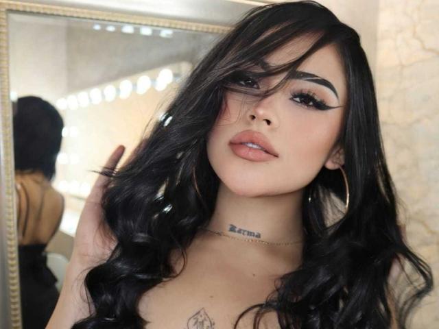 LilitthCorper has long, wavy black hair, full lips, and thick eyebrows. She's wearing large hoop earrings and has a tattoo on her neck. Her tits are visible.
