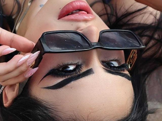LilitthCorper's face, black eyebrows shaped into thick, angry lines, wearing black sunglasses, pink glossy lips, long pink nails, black hair, holding sunglasses with her right hand.