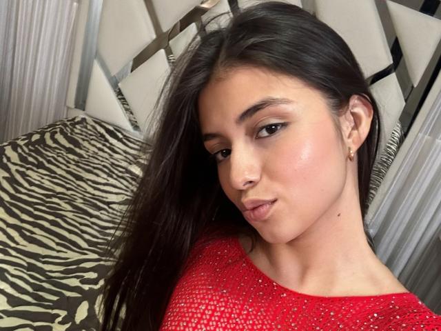 IsabelaRios with long dark hair, wearing a red sparkly top, small gold earrings, pouty lips, and a slight smile, showing her smooth, shiny face. Zebra-patterned pillow in background.