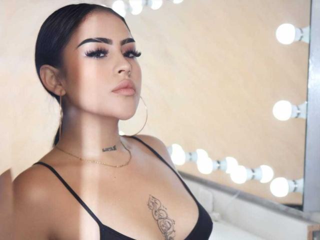 LilitthCorper with dark hair, hoop earrings, black top, tattooed chest, and eyeliner, looking smug. Boobs visible, small gold necklace, light skin, and long eyelashes.