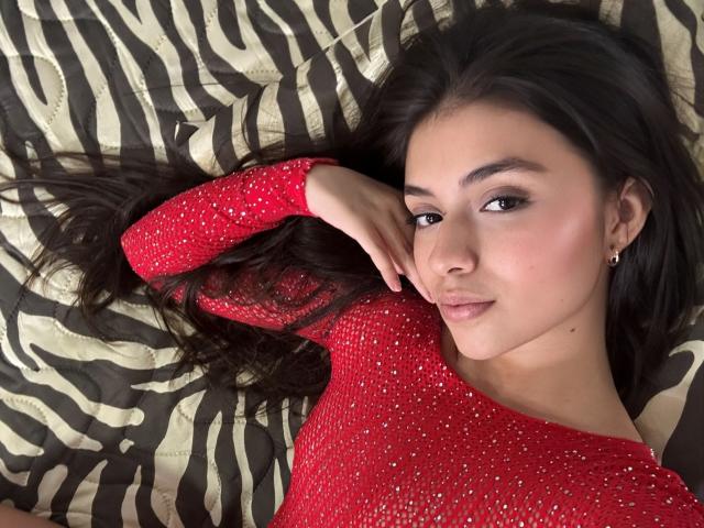 IsabelaRios with long black hair, wearing a red sparkly top, lying on zebra-patterned bed, looking at camera with a slight smile, hand touching her face.