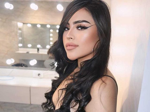 LilitthCorper with long black wavy hair, thick eyebrows, eyeliner, full pink lips, and hoop earrings, wearing a black top, in a bathroom with lit mirrors.