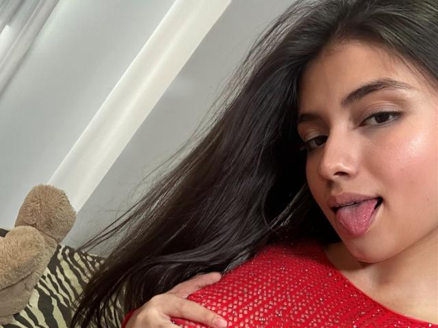 IsabelaRios with long dark hair, brown eyes, and slightly parted lips sticking out her tongue. She's wearing a red, mesh top, and has a beige stuffed toy on the left.