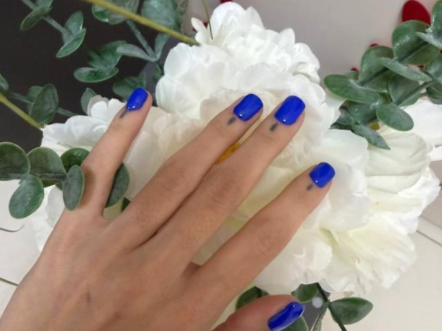 MoonLiza's hand with bright blue nails, tattoos on fingers, resting on white flower. Green leaves in background. Soft, natural skin tone. Handsome, feminine. Fluffy petals.