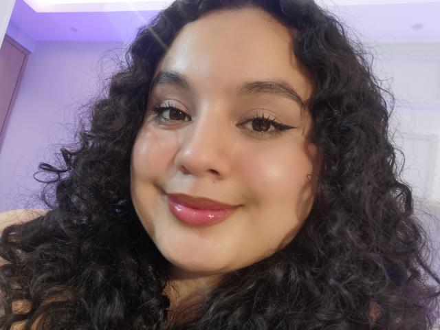 TaniaRichmond has curly black hair, brown eyes, and glossy pink lips. She's smiling with a small nose piercing and eyeliner. Her skin is light brown and she's looking at the camera.