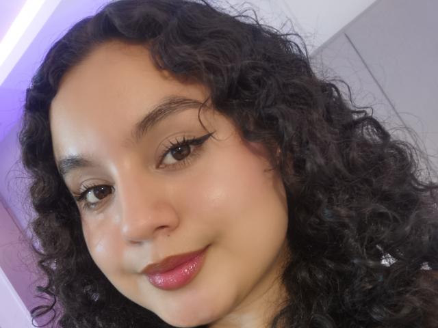TaniaRichmond has curly black hair, brown eyes with long eyelashes, pink glossy lips, and a light brown skin tone. She's smiling softly at the camera.