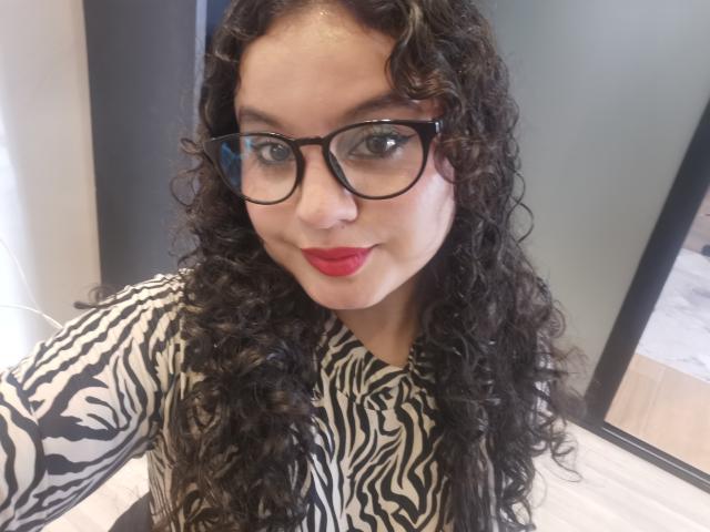 TaniaRichmond with curly black hair, wearing zebra-patterned shirt, black glasses, and red lipstick, smiling at the camera. Boobs are not visible. She's indoors, looking confident.