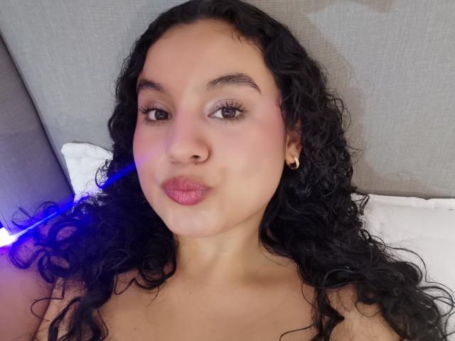 TaniaRichmond has curly black hair, brown eyes, pink lipstick, and gold earrings. She's looking at the camera with a slight pout. Her tits are not visible.