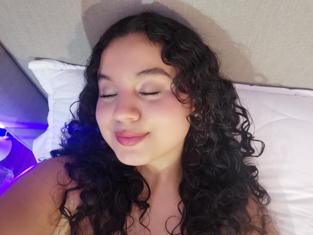 TaniaRichmond with curly black hair, pink eyelids, and pink lips, sleeping on white pillows, with a purple light on the left side of the image.