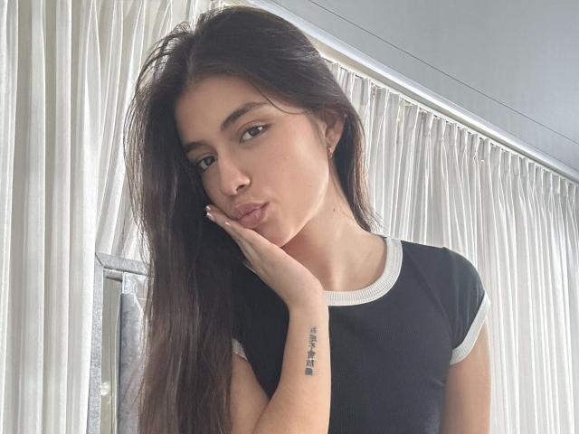 IsabelaRios with long dark hair, pouty lips, hand on cheek, black and white tee, tattoo on right wrist, standing in front of white curtains, looking at camera.