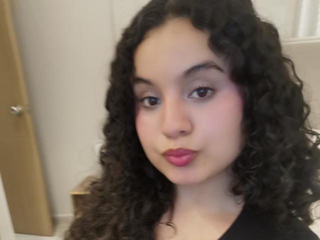 TaniaRichmond has curly black hair, brown eyes, pink lipstick, and a light skin tone. She's wearing a black shirt, looking at the camera with a slight pout.