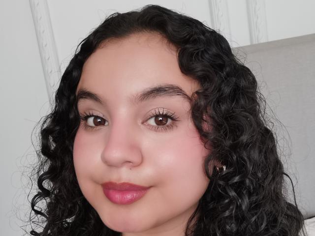 TaniaRichmond's face with curly black hair, pink lipstick, and brown eyes, showing a slight smile. Her skin is light brown with a pinkish blush. Her eyelashes are long.