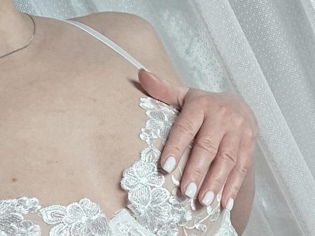 GladisBelle's hand with white nails rests on her lace-covered boobs, showing her light skin and delicate lace bra with floral patterns. Soft light highlights her gentle touch.