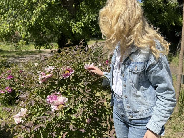 GladisBelle mit blonden, gewellten Haaren, einer Jeansjacke und blauen Jeans, die rosa Rosen an einem grünen Busch berührt, steht draußen an einem sonnigen Tag..