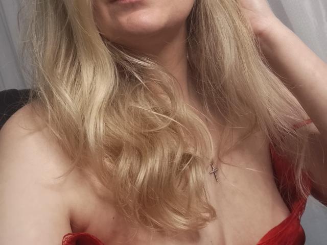 GladisBelle's blonde hair, wavy and flowing, covers her tits, wearing a red top, her hand in her hair, a small cross necklace, soft light on her fair skin.