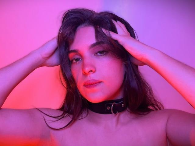 Katarina Novak, les mains dans ses cheveux foncés, portant un collier noir avec une boucle en argent, sous une lumière rose et violette. Elle a une expression neutre et est nue..