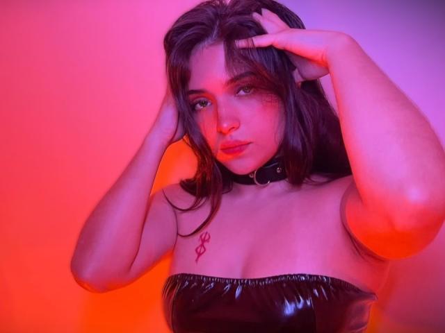 Katarina Novak, avec ses cheveux foncés, les mains dans les cheveux, portant un haut noir brillant sans bretelles, un col, et un tatouage rouge sur la poitrine, sous un éclairage rose et orange..