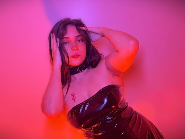 KatarinaNovak with wet black latex dress, choker, and tattooed Ankh on chest, hand in dark hair, pink and red lighting. Her tits are medium, and her ass is hidden.