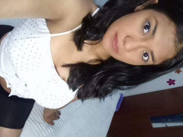 ArianithaXox has medium brown skin, black wavy hair, and is wearing a white polka dot top with black shorts, showing small tits and a flat ass.