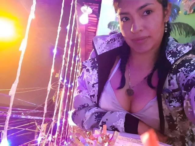 ArianithaXox with tan skin, medium tits, black bra, and white patterned jacket, wearing a necklace, has braids, and a slight smile, under colorful string lights.