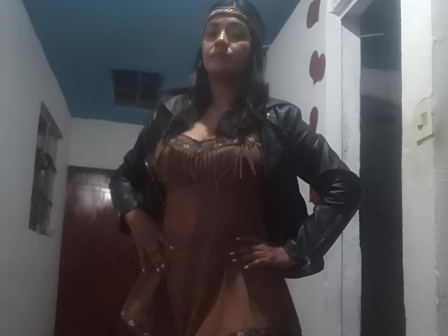 ArianithaXox with hands on hips, wearing a brown dress with fringe, black leather jacket, and headband, standing in a dimly lit room, showing her boobs.