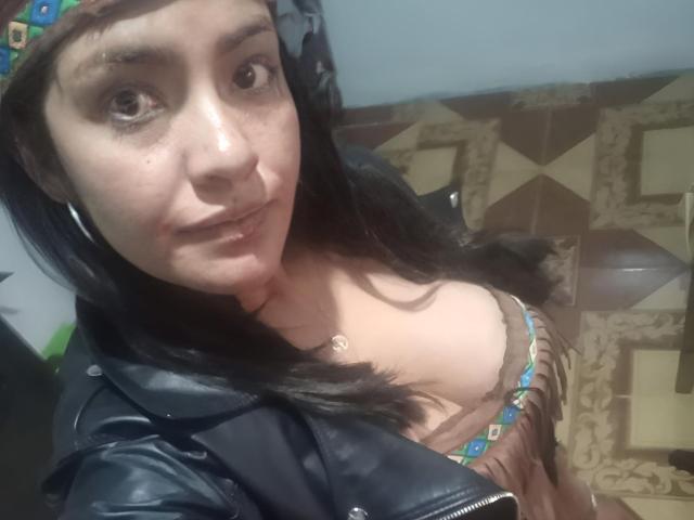 ArianithaXox with brown eyes, wearing a leather jacket, headband, and frilled dress showing her tits, standing in a room with patterned floor. She has a slight smile.