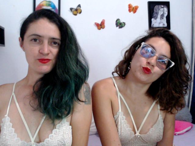 Due donne LunaVenus con il seno scoperto, vestite con lingerie di pizzo bianco, una con capelli verdi, rossetto rosso e tatuaggi, l'altra con capelli castani ricci, occhiali e rossetto rosso, che si baciano..