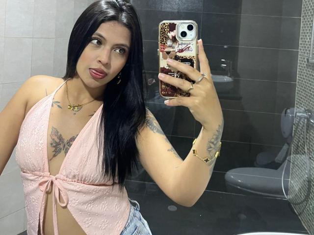 NyraVanel, avec ses cheveux noirs, porte un haut rose à lacets qui dévoile sa poitrine, et elle a des tatouages sur la poitrine et le bras. Elle prend un selfie devant un miroir avec une coque de téléphone à motifs léopard. Elle porte du rouge à lèvres..