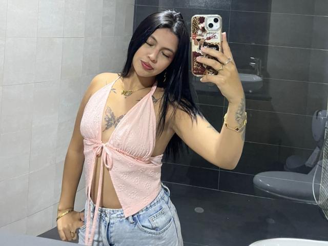 NyraVanel, avec ses longs cheveux noirs, un haut bandeau rose qui révèle ses seins, un jean bleu, des tatouages sur les bras, tenant un téléphone avec une coque à motifs floraux, debout dans une salle de bain, se prenant en photo devant le miroir..