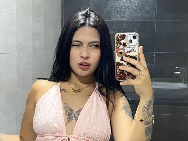 NyraVanel, avec de longs cheveux noirs, une robe en dentelle rose, des tatouages sur la poitrine et les bras, la langue tirée, tenant un téléphone avec une coque à motifs léopard, dans un selfie devant un miroir de salle de bain..