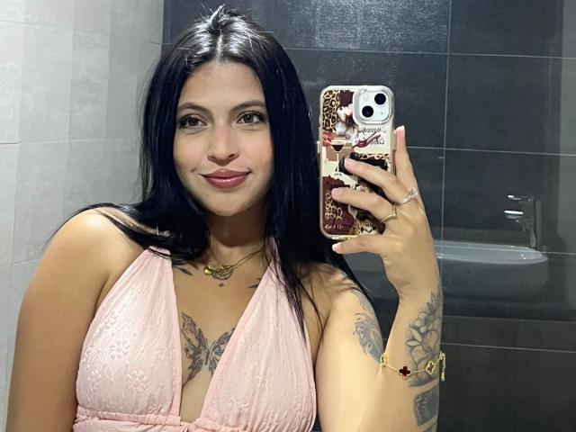 NyraVanel with black hair, pink lace dress, tattoos on arms, holding phone with leopard print case, smiling, pink lipstick, gold necklace, and ring, in bathroom mirror selfie.