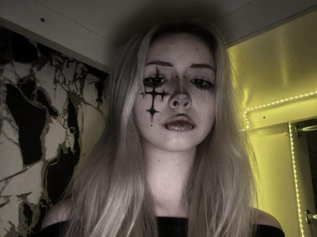 AmeliaCarratura with black star-shaped makeup on her left cheek, pale skin, blonde hair, and a slightly open mouth, wearing an off-shoulder black top, under dim yellow lighting.