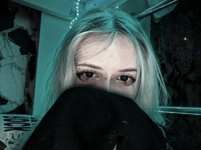 AmeliaCarratura's pale face with blonde hair, black eyeliner, and moles, peeks from under black clothes, eyes looking up, surrounded by blue-lit metal bars.