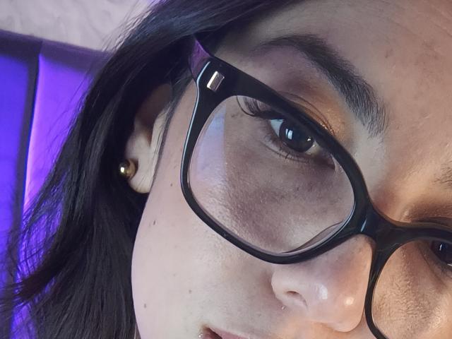 MiaReed's face with dark glasses, black hair, gold ear stud, and brown eyes, showing a slight smile, with visible skin texture and subtle makeup.